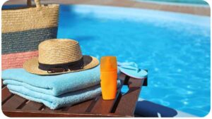 towels, hat, sunglasses and a straw hat sit on a wooden table near a swimming pool